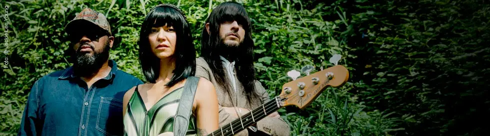 Drei Personen stehen vor einer grünen, bewachsenen Wand, eine Frau in der Mitte hält einen E-Bass.