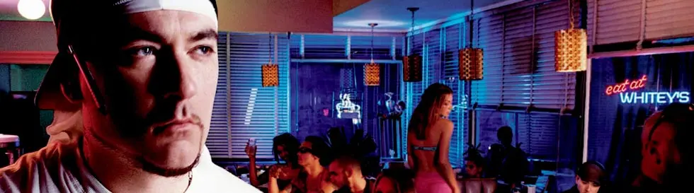 Eine Person mit weißem Stirnband, ein Kuli hinterm Ohr in einem belebten, farbig beleuchteten Raum mit Gästen im Hintergrund.