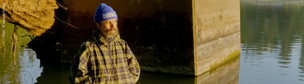 Eine Person mit Brille trägt eine blaue Mütze und karierte Kleidung vor einem dunklen Hintergrund.