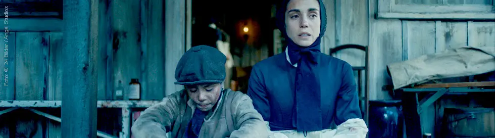 Zwei Personen sitzen vor einer Holzhütte; links ein Junge, rechts eine Frau mit Kopftuch, beide in einfachen Kleidern.