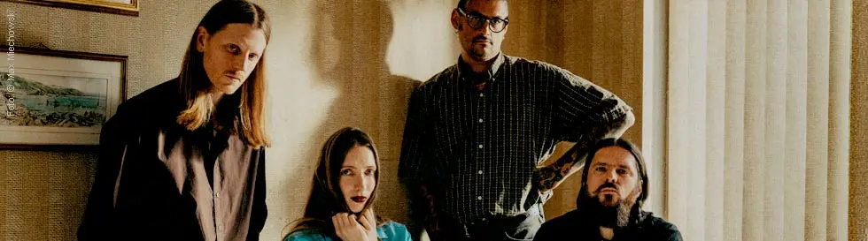 Vier Personen in einem Raum mit beigen Wänden. Eine Frau in türkisfarbener Kleidung sitzt, während drei Männer stehen.