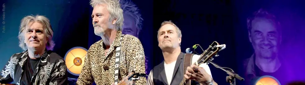 Drei Männer spielen Gitarre auf einer Bühne, sie tragen lässige Kleidung. Im Hintergrund sind bunte Lichter.