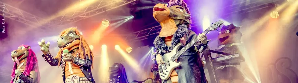 Fünf Dinosaurier in Rock-Outfits spielen auf einer Bühne mit Instrumenten unter bunten Lichtern.