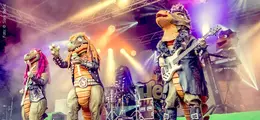 Fünf Dinosaurier in Rock-Outfits spielen auf einer Bühne mit Instrumenten unter bunten Lichtern.