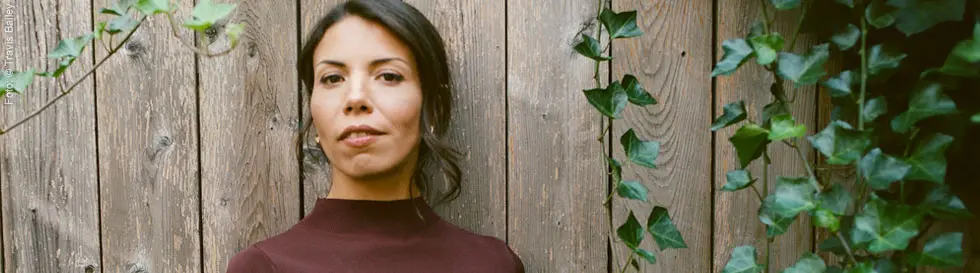 Frau mit dunklem Haar vor Holzwand, Efeu rankt seitlich.