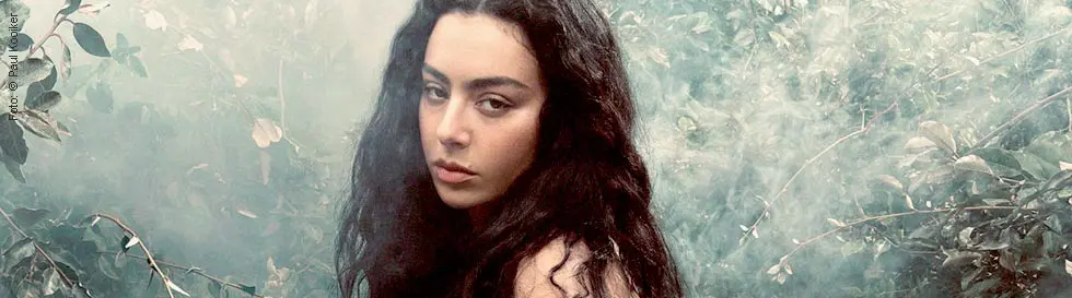 Frau mit langen, dunklen Haaren sitzt vor nebligem, blättrigem Hintergrund und schaut ernst zur Seite.