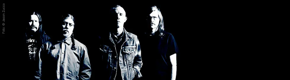Die Aufnahme zeigt vier Männer in schwarz-weißer Bearbeitung vor dunklem Hintergrund. Links ist ein Fotokredit.