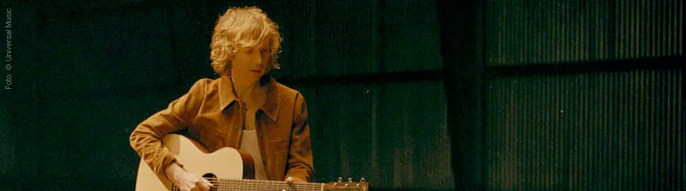 Eine Person mit lockigem Haar spielt Gitarre und trägt eine braune Jacke in einem dunklen Raum.