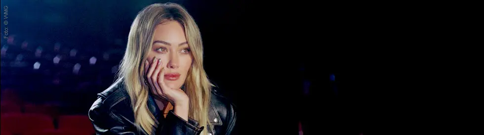 Frau in schwarzer Lederjacke, nachdenklich, stützt ihren Kopf auf die Hand, sitzt vor unscharfem Hintergrund.