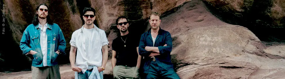 Vier Männer stehen vor einem Felsen. Sie tragen lässige Kleidung und Sonnenbrillen.