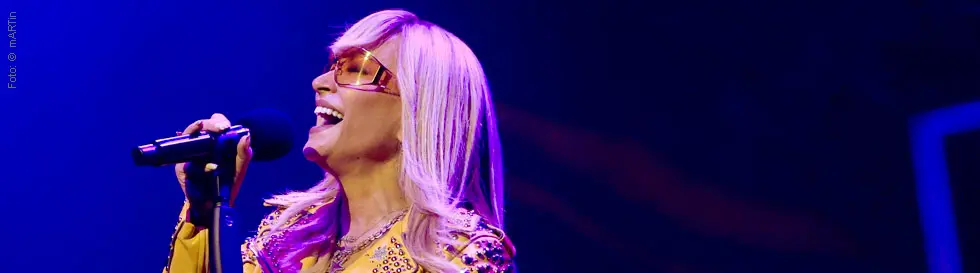 Eine Frau singt leidenschaftlich in ein Mikrofon, trägt eine gelbe Jacke mit Nieten und eine große Brille.