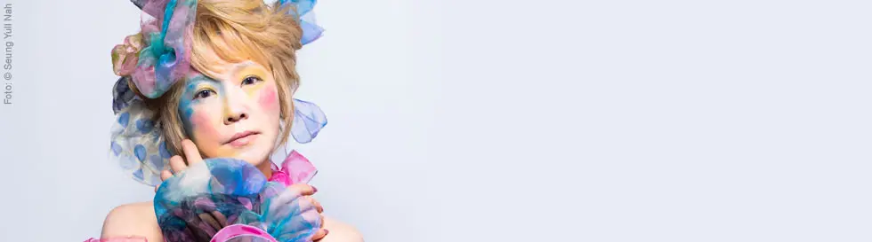 Person mit buntem Make-up und farbenfrohem Stoff im Haar; pastellfarbene Töne dominieren das Gesicht.