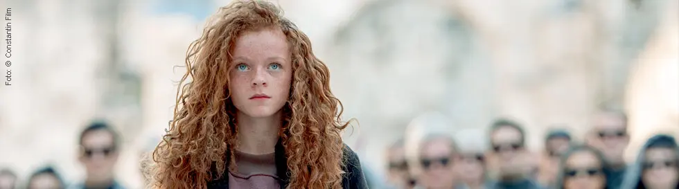 Ein Mädchen mit lockigem, rotem Haar steht im Vordergrund, umgeben von einer Gruppe in grauer Kleidung im Hintergrund.