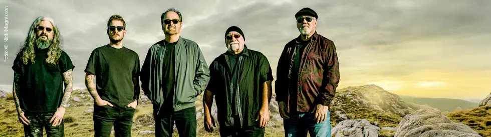 Eine Gruppe von fünf Männern steht nebeneinander auf einer grasbewachsenen, felsigen Landschaft.