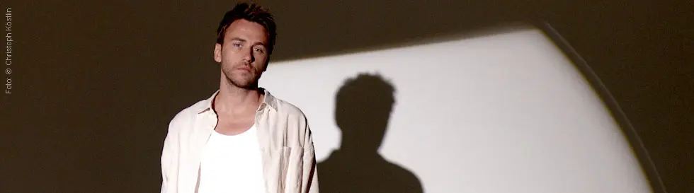 Person in weißem Hemd und T-Shirt, Hände in den Hosentaschen, steht vor neutralem Hintergrund mit Schatten.