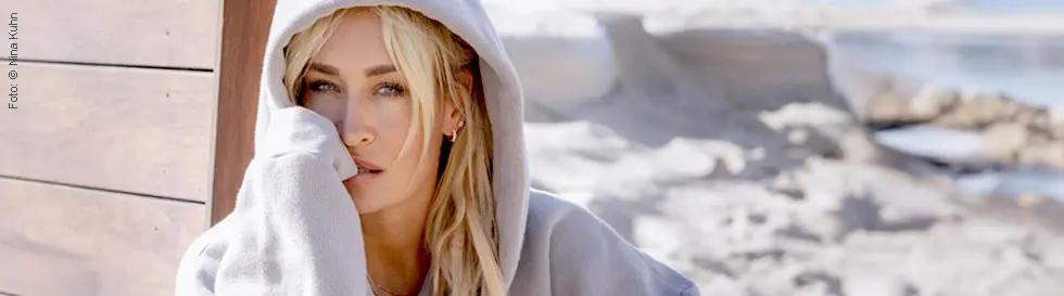 Eine blonde Person in einem grauen Hoodie sitzt entspannt am Strand, gestützt gegen eine Holzwand.