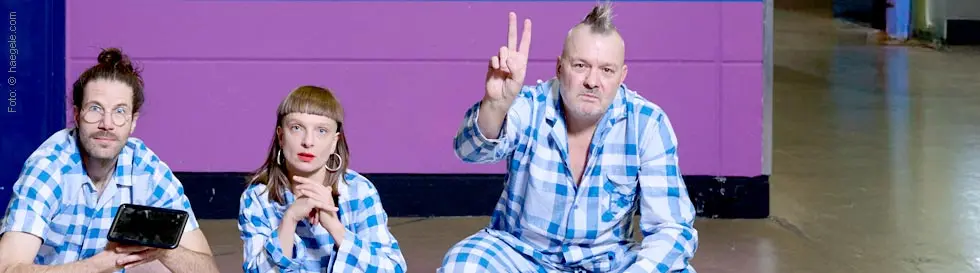 Drei Personen in blau-weiß karierten Anzügen hocken nebeneinander, eine hält ein Gerät, eine macht ein Peace-Zeichen.