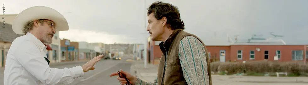 Ein weiß gekleideter Mann mit Cowboyhut und Sheriffstern spricht gestikulierend mit einem Mann in brauner Weste auf der Straße.