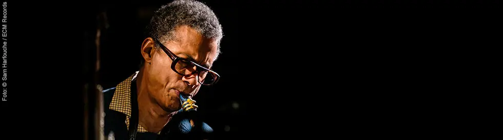 Ein Mann mit Brille spielt Saxophon im Halbdunkel. Er trägt ein kariertes Hemd unter einem dunklen Pullover.