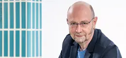 Ein Mann mit Brille und Bart sitzt lächelnd im Vordergrund, im Hintergrund vertikale blaue Streifen.