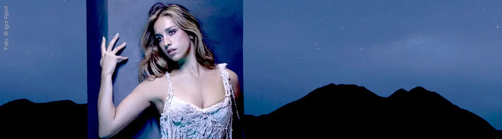 Eine Frau in weißem Kleid lehnt an einer Wand, umgeben von einem nächtlichen, bläulichen Hintergrund mit Hügeln.