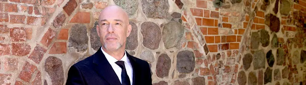 Ein Mann in einem Anzug steht mit einem Koffer vor einer alten Steinmauer.