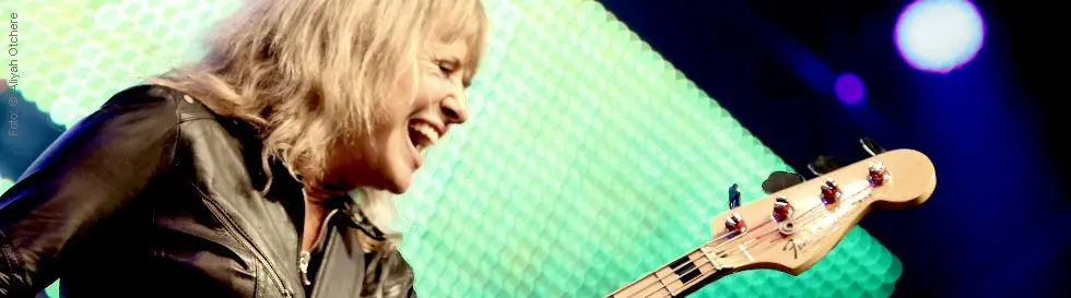 Eine Frau spielt lächelnd Bassgitarre auf einer Bühne, beleuchtet von buntem Licht.