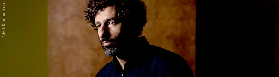 Mann mit lockigem Haar und Vollbart, in dunkler Kleidung, steht vor einem braunen Hintergrund mit Schattenwirkung.