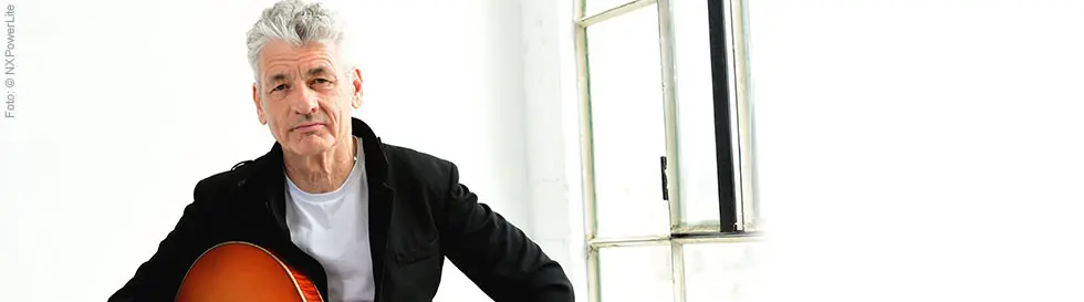 Ein Mann mit grauen Haaren hält eine Gitarre, sitzt vor einem hellen Fenster und trägt ein schwarzes Sakko über einem weißen Shirt.