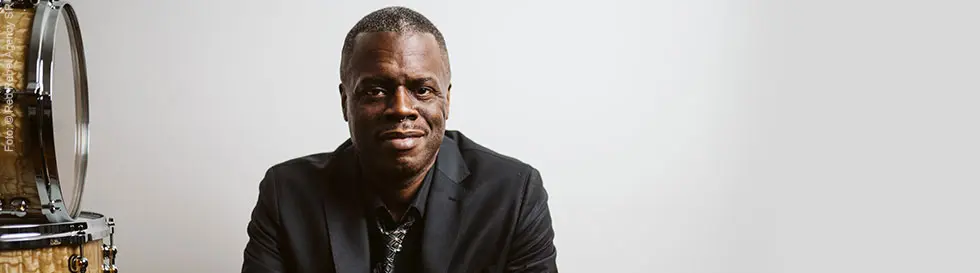 Der Mann trägt einen dunklen Anzug und sitzt entspannt neben einem Schlagzeug. Er lächelt leicht und trägt eine gemusterte Krawatte.