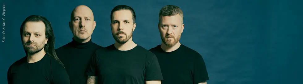 Vier Männer in schwarzen T-Shirts stehen vor einem blauen Hintergrund, sie schauen direkt nach vorne.