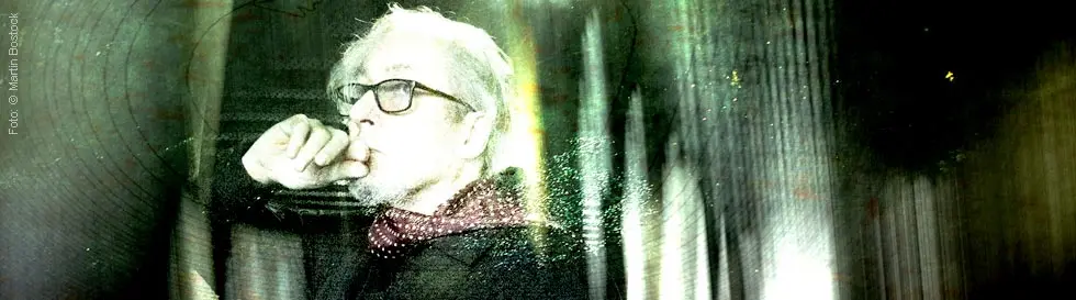 Eine ältere Person mit Brille und Schal denkt nach, Hand am Kinn. Die Szenerie wirkt künstlerisch bearbeitet.
