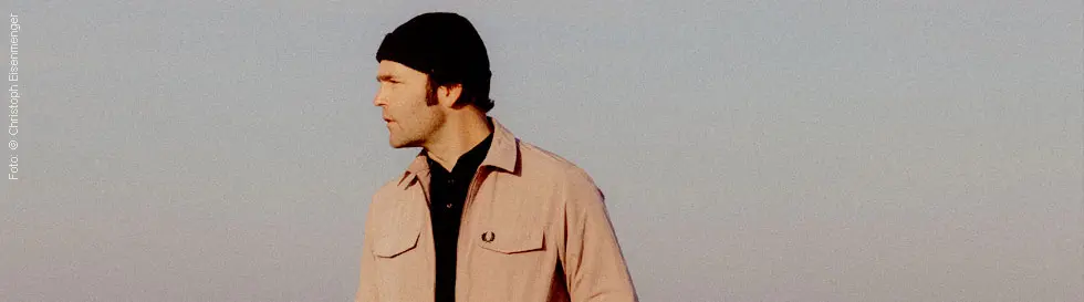 Ein Mann trägt eine Mütze und schaut zur Seite. Er trägt eine helle Jacke mit einem kleinen Logo auf der Brust.