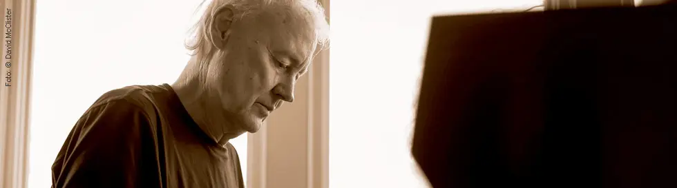 Ein nachdenklicher älterer Mann mit hellem Haar in einem dunklen T-Shirt, sitzt an einem Klavier.