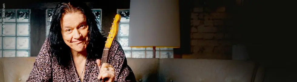 Eine Person sitzt auf einem Sofa und hält eine E-Gitarre. Im Hintergrund steht eine Lampe.