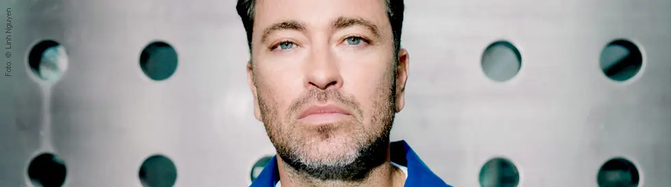 Ein Mann mit Bart und blauen Augen, vor einer metallischen, gelochten Wand.