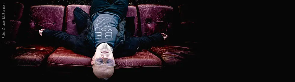Person liegt auf einem alten, roten Sofa. Kleidung kontrastiert mit weißem Gesicht.