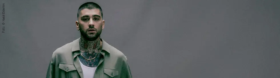 Ein Mann mit kurzem Haar, Tattoo am Hals, in grüner Jacke. Sein Gesichtsausdruck ist ernst.