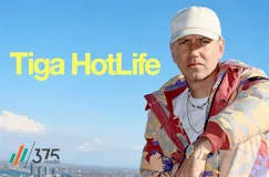 Gelber Text: "Tiga HotLife". Eine Person in bunter Jacke und Mütze mit städtischem Hintergrund.
