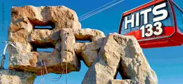 Große Steinskulptur mit den Buchstaben "BRAVO". Luftseilbahnwagen mit der Aufschrift "HITS 133" vor blauem Himmel.