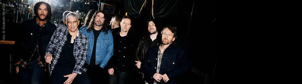 Sechs Männer in lässiger Kleidung stehen in einer Bar; einer trägt eine karierte Jacke. Die Atmosphäre ist entspannt.