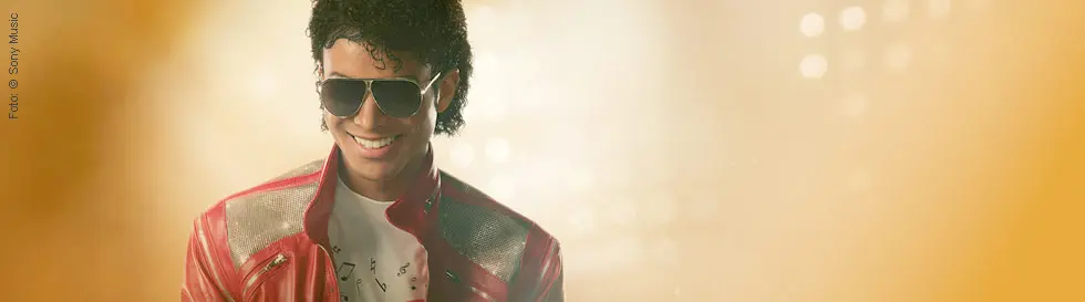 Ein Mann mit lockigem Haar trägt eine Sonnenbrille und rote Jacke. Helles, unscharfes Licht im Hintergrund.