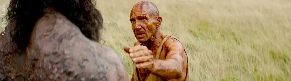 Zwei schmutzige, erschöpfte Männer in zerlumpter Kleidung stehen auf einem trockenen Feld, einer streckt die Hand aus.