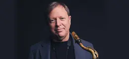 Ein Mann in dunkler Kleidung hält ein Saxophon und lächelt leicht vor einem dunklen Hintergrund.