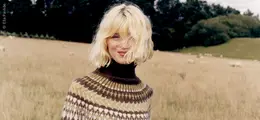 Eine Person mit blondem Haar steht auf einer Wiese, trägt einen gemusterten Pullover. Im Hintergrund sind Bäume und Schafe.