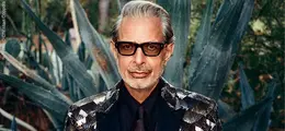 Ein Mann mit Brille und glamourösem Anzug in einer Gartenlandschaft.