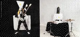 Links posiert eine Figur mit Gitarre, rechts sitzt eine Person an einem Schlagzeug. Beide tragen gepunktete Kostüme.