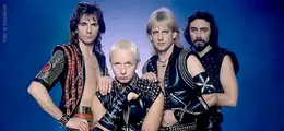 Vier Männer in rockiger Lederkleidung vor blauem Hintergrund. Sie blicken ernst und tragen Nieten und Metallaccessoires.