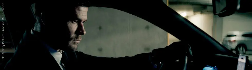 Ein Mann in einem Anzug fährt ein Auto in einer Tiefgarage, das Armaturenbrett leuchtet blau.