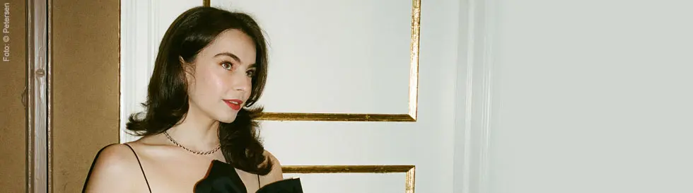 Frau mit dunklem Haar in elegantem schwarzen Kleid, vor einer klassisch dekorierten Wand.